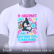Футболка лаборант білого кольору, складена на столі, фон світло-сірий, принт з мікроскопом, пробірками та текстом, прикріплений етикеткою з зеленим шнурком.