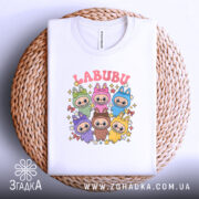 Футболка labubu білого кольору, складена на плетеній кошику, фон світлий, принт з кольоровими персонажами та написом 'LABUBU' у верхній частині.