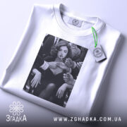 Футболка леді у центрі уваги, біла, зображена зверху, на фоні світлого градієнта, принт з чорно-білим зображенням жінки з сигаретою, з етикеткою на комірці.