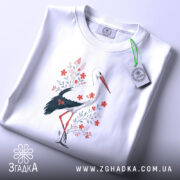 Футболка лелека з квітами, білого кольору, складена на поверхні, фон світло-сірий, принт з лелекою та червоними квітами, з етикеткою на комірці.
