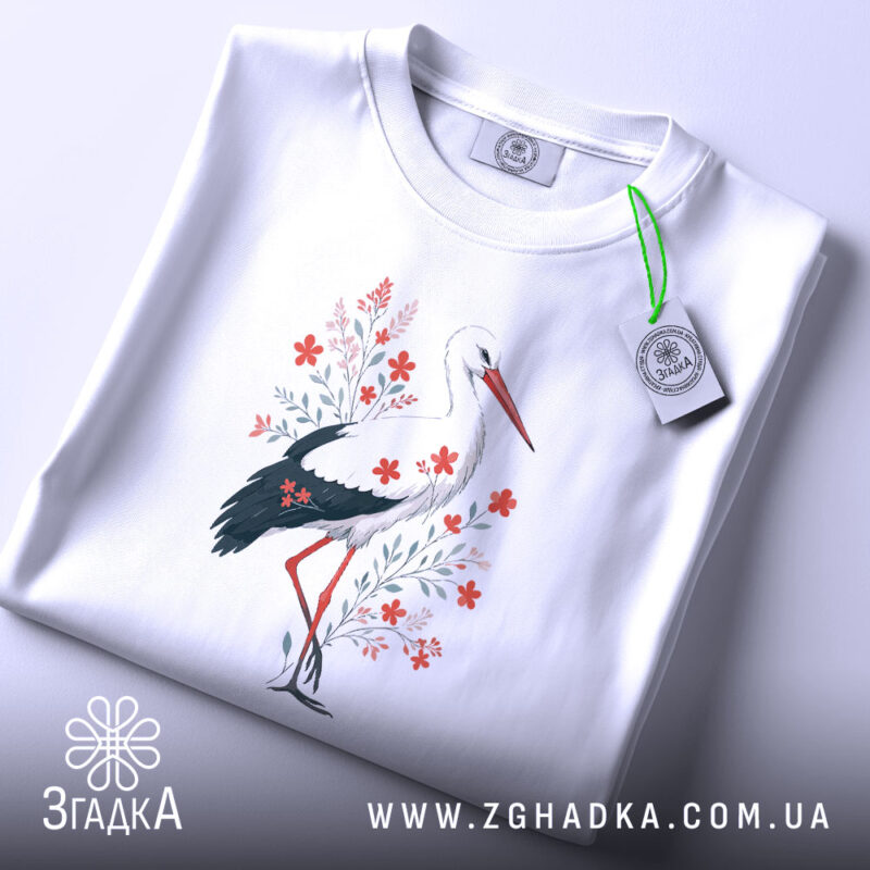 Футболка лелека з квітами, білого кольору, складена на поверхні, фон світло-сірий, принт з лелекою та червоними квітами, з етикеткою на комірці.