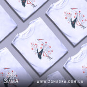 Футболка лелека з квітами, білого кольору, зображена з ракурсу спереду на манекені, фон світло-сірий, принт з лелекою та червоними квітами на зеленому листі.