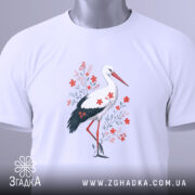 Футболка лелека з квітами, білого кольору, складена на поверхні, фон світло-сірий, принт з лелекою та червоними квітами, з етикеткою на комірці.