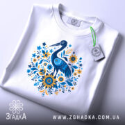 Футболка лелека з квітами, біла, зображена зверху, фон світло-сірий, принт з лелекою та квітами в блакитних і жовтих тонах, з етикеткою на зеленому шнурку.