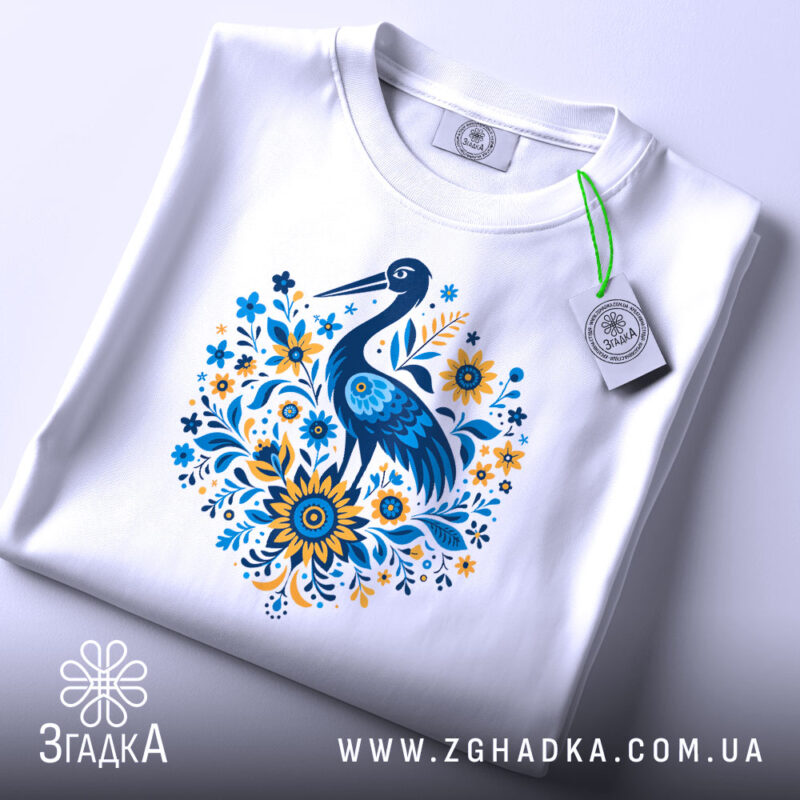 Футболка лелека з квітами, біла, зображена зверху, фон світло-сірий, принт з лелекою та квітами в блакитних і жовтих тонах, з етикеткою на зеленому шнурку.