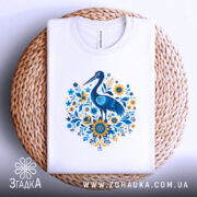 Футболка лелека з квітами, біла, зображена зверху на плетеній основі, фон світло-сірий, принт з лелекою та квітами в блакитних і жовтих тонах.