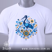 Футболка лелека з квітами, біла, зображена зверху, фон світло-сірий, принт з лелекою та квітами в блакитних і жовтих тонах, з етикеткою на зеленому шнурку.