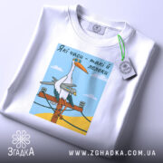 Футболка лелеки воєнного часу, біла, складена на плоскій поверхні, фон світло-сірий, принт з лелекою на електричному стовпі, прикрашена етикеткою з зеленим шнурком.