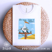 Футболка лелеки воєнного часу, біла, складена на плоскій поверхні з плетеним кошиком, фон світло-сірий, принт з лелекою на електричному стовпі, деталі принта чітко видимі.