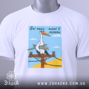 Футболка лелеки воєнного часу, біла, складена на плоскій поверхні, фон світло-сірий, принт з лелекою на електричному стовпі, прикрашена етикеткою з зеленим шнурком.