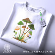 Футболка лісові магічні гриби, біла, складена на поверхні, фон світло-сірий, принт з грибами, прикрашений етикеткою з зеленим шнурком.