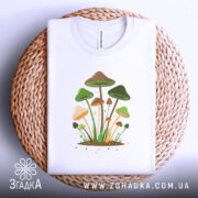 Футболка лісові магічні гриби, біла, складена на кошику з плетеного матеріалу, фон білий, принт з грибами, деталі зеленого та коричневого кольору.