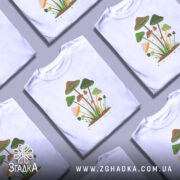 Футболка лісові магічні гриби, біла, зображена на манекені, фон світло-сірий, принт з різнокольоровими грибами на зеленому фоні з зірочками.