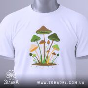 Футболка лісові магічні гриби, біла, складена на поверхні, фон світло-сірий, принт з грибами, прикрашений етикеткою з зеленим шнурком.