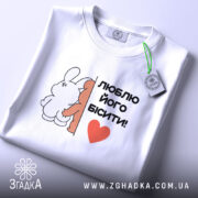 Футболка люблю його бісити, біла, складена на столі, фон світло-сірий, принт з двома персонажами, текст чорний з червоним сердечком, на комірці є етикетка з зеленим шнурком.