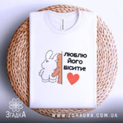 Футболка люблю його бісити, біла, складена на плетеній кошику, фон світло-сірий, принт з двома персонажами, текст чорний з червоним сердечком.