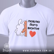 Футболка люблю його бісити, біла, складена на столі, фон світло-сірий, принт з двома персонажами, текст чорний з червоним сердечком, на комірці є етикетка з зеленим шнурком.