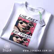 Футболка Live Fast Eat Trash білого кольору, складена на столі, ракурс зверху, фон світло-сірий, принт з двома тваринами в окулярах, текст на футболці чорний і рожевий, на футболці є етикетка з зеленим шнурком.
