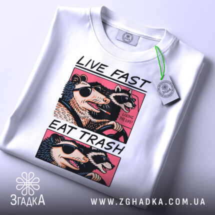 Футболка Live Fast Eat Trash білого кольору, складена на столі, ракурс зверху, фон світло-сірий, принт з двома тваринами в окулярах, текст на футболці чорний і рожевий, на футболці є етикетка з зеленим шнурком.