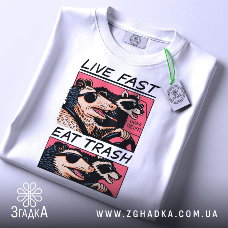 Футболка Live Fast Eat Trash білого кольору, складена на столі, ракурс зверху, фон світло-сірий, принт з двома тваринами в окулярах, текст на футболці чорний і рожевий, на футболці є етикетка з зеленим шнурком.