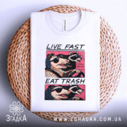 Футболка Live Fast Eat Trash білого кольору, складена на плетеній основі, ракурс зверху, фон світло-сірий, принт з двома тваринами в окулярах, текст на футболці чорний і рожевий.