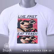 Футболка Live Fast Eat Trash білого кольору, складена на столі, ракурс зверху, фон світло-сірий, принт з двома тваринами в окулярах, текст на футболці чорний і рожевий, на футболці є етикетка з зеленим шнурком.