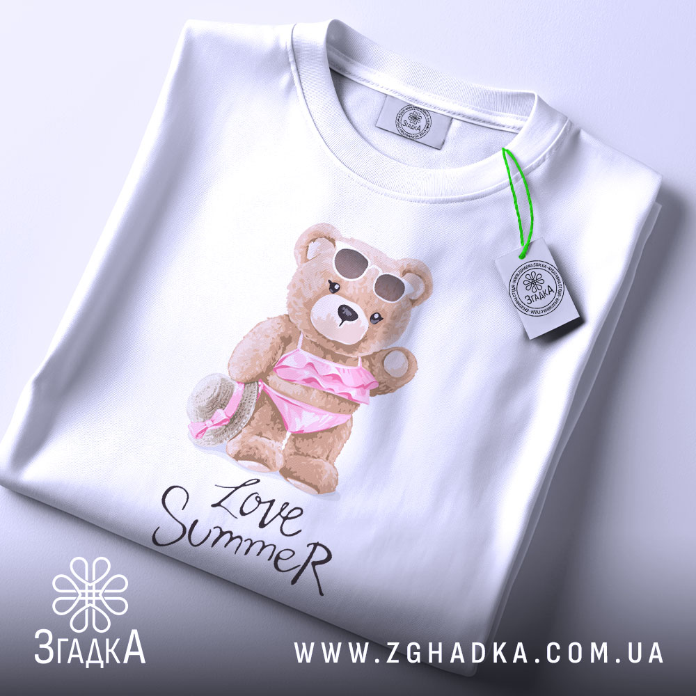 Футболка Love Summer Bear, біла, складена на столі, фон світлий, принт з ведмедиком у купальнику з рожевими рюшами та сонцезахисними окулярами, на футболці є етикетка з логотипом.