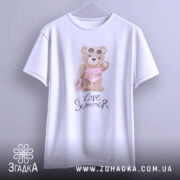 Футболка Love Summer Bear, біла, підвішена на вішалці, фон нейтральний, принт з ведмедиком у купальнику з рожевими рюшами та сонцезахисними окулярами, напис 'Love Summer' під принтом.