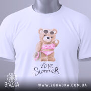 Футболка Love Summer Bear, біла, складена на столі, фон світлий, принт з ведмедиком у купальнику з рожевими рюшами та сонцезахисними окулярами, на футболці є етикетка з логотипом.