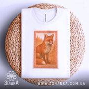 Футболка лисиця білого кольору, складена на плетеному кошику, з верхнього ракурсу. Принт зображає лисицю в оранжевих тонах, оформлений в рамку, на фоні світло-сірого кольору.