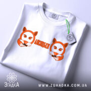 Футболка лисицьки білого кольору, складена на поверхні, з видимим етикеткою, фон світлий, принт з двома лисицями та написом 'ЛИСИЦЬКИ' оранжевого кольору.