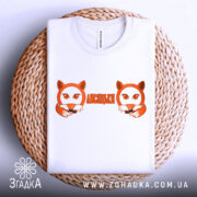 Футболка лисицьки білого кольору, складена на плетеній основі, фон світлий, принт з двома лисицями та написом 'ЛИСИЦЬКИ' оранжевого кольору.