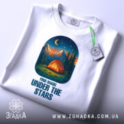 Футболка магія нічного лісу, біла, зображена зверху, на фоні світло-сірого градієнта, принт з малюнком намету, гір та зірок на нічному небі, з етикеткою на рукаві.