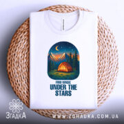 Футболка магія нічного лісу, біла, зображена зверху, на фоні плетеного кошика, принт з малюнком намету, гір та зірок на нічному небі.