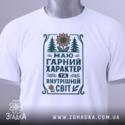 Футболка маю гарний характер. Біла футболка, складена на поверхні, з видимим принтом. На фоні світло-сірий градієнт. Принт містить текст та малюнки, включаючи соняшник і дерева, виконані в зелених і жовтих тонах, а також етикетка з зеленим шнурком.