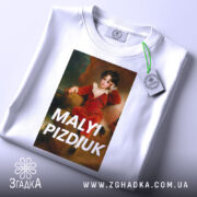 Футболка малий шибеник, біла, складена на столі, фон світло-блакитний, принт з портретом хлопчика в червоному костюмі, текст 'MALYI PIZDIUK' великими літерами, на футболці є етикетка з зеленим шнурком.
