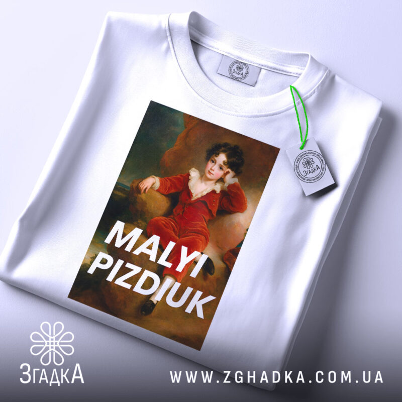 Футболка малий шибеник, біла, складена на столі, фон світло-блакитний, принт з портретом хлопчика в червоному костюмі, текст 'MALYI PIZDIUK' великими літерами, на футболці є етикетка з зеленим шнурком.
