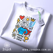 Футболка мама наша сила, білого кольору, складена на поверхні, фон світло-сірий, принт зображує супергероїню з червоним плащем, синім верхом, оточену серцем, сонцем, квітами та трофеєм, на футболці є етикетка.