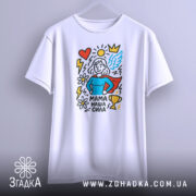 Футболка мама наша сила, білого кольору, підвішена на вішалці, фон світло-сірий, принт зображує супергероїню з червоним плащем, синім верхом, серцем, сонцем, квітами та трофеєм.