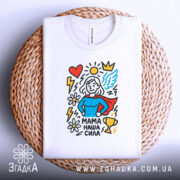 Футболка мама наша сила, білого кольору, складена на плетеній основі, фон білий, принт містить малюнок супергероїні з червоним плащем, синім верхом, оточену серцем, сонцем, квітами та трофеєм.