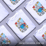 Футболка мама наша сила, білого кольору, зображена на манекені, фон світло-сірий, принт містить малюнок супергероїні з червоним плащем, синім верхом, серцем, сонцем, квітами та трофеєм.