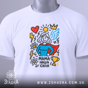 Футболка мама наша сила, білого кольору, складена на поверхні, фон світло-сірий, принт зображує супергероїню з червоним плащем, синім верхом, оточену серцем, сонцем, квітами та трофеєм, на футболці є етикетка.