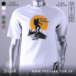 Футболка мандрівки надихають. Біла футболка з принтом, на якому зображений альпініст на вершині гори на фоні жовтого сонця. Принт виконаний у чорному кольорі, з деталями лісу та гір. Футболка представлена на манекені з блискучою поверхнею.