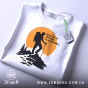 Футболка мандрівки надихають. Біла футболка, складена на поверхні, з принтом альпініста на фоні жовтого сонця. Принт чорного кольору, деталі лісу та гір чітко видимі. На футболці є етикетка з зеленим шнурком.