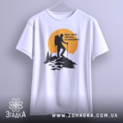 Футболка мандрівки надихають. Біла футболка, підвішена на вішалці, з принтом альпініста на фоні жовтого сонця. Принт чорного кольору, з деталями лісу та гір, виглядає стильно та сучасно.
