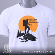 Футболка мандрівки надихають. Біла футболка, складена на поверхні, з принтом альпініста на фоні жовтого сонця. Принт чорного кольору, деталі лісу та гір чітко видимі. На футболці є етикетка з зеленим шнурком.