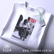 Футболка мавпа на велосипеді, біла, складена на поверхні, фон світлий, принт зображає мавпу на велосипеді з написом 'LIFE', на футболці є етикетка.