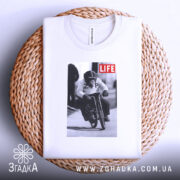 Футболка мавпа на велосипеді, біла, складена на плетеній основі, фон світлий, принт зображає мавпу на велосипеді з написом 'LIFE'.