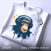 Футболка мавпа в навушниках, біла, зображена на столі з невеликим ракурсом зверху, фон світло-сірий, принт зображає мавпу в навушниках з кольоровими плямами та метеликами, з етикеткою на рукаві.