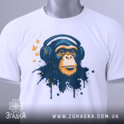 Футболка мавпа в навушниках, біла, зображена на столі з невеликим ракурсом зверху, фон світло-сірий, принт зображає мавпу в навушниках з кольоровими плямами та метеликами, з етикеткою на рукаві.